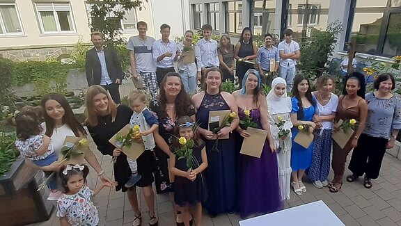 Eine Gruppe von Menschen steht in einem Garten, viele halten Blumen und Urkunden in den Händen. Einige Kinder sind ebenfalls dabei. Es scheint eine Feier zu sein, möglicherweise zur Ehrung oder Graduierung. Im Hintergrund sind Pflanzen und Gebäude sichtbar.
