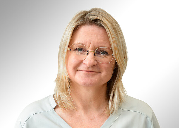 Porträtfoto einer lächelnden Frau mittleren Alters mit schulterlangen blonden Haaren und Brille vor hellem, neutralem Hintergrund. Sie trägt eine helle Bluse und blickt direkt in die Kamera.