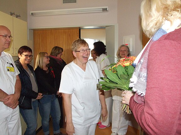 Gruppe von Krankenhausmitarbeitenden in einem Flur. Eine Pflegekraft in Weiß lächelt überrascht, während ihr jemand einen bunten Blumenstrauß überreicht. Die anderen schauen zu und freuen sich – eine kleine Feier oder Verabschiedung.