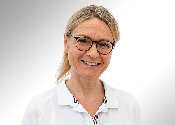 Porträt einer lächelnden Frau mit blondem, zurückgebundenem Haar und runden Brillen. Sie trägt ein weißes Poloshirt und blickt direkt in die Kamera vor neutralem, hellgrauem Hintergrund.