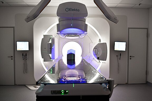 Strahlentherapiegerät in Klinik, Blick frontal entlang Behandlungsliege zum Linearbeschleuniger (Elekta), blaue Beleuchtung, runder Gantry-Ring; Monitore an den Seiten. Leerer Raum, bereit für Patientenbehandlung gegen Krebs.