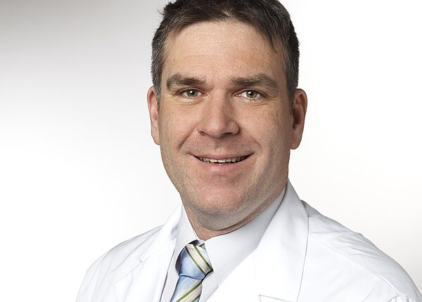 Porträt eines lächelnden Mannes in weißem Laborkittel und gestreifter Krawatte, mit kurz geschnittenen dunklen Haaren vor neutralem Hintergrund; Schwerpunkt auf seinem freundlich-professionellen Auftreten.