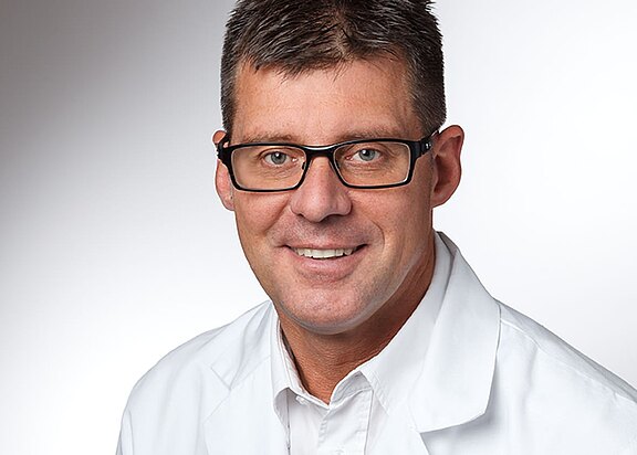 Porträt eines lächelnden Mannes mit kurzer dunkler Frisur und schwarzer Brille, in weißem Laborkittel, vor neutralem Hintergrund. Nahaufnahme von Kopf und Oberkörper; professionelles Profilfoto.