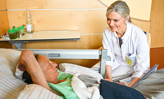 Eine Ärztin sitzt am Bett eines älteren Patienten im Krankenzimmer. Sie lächelt, hält eine Mappe mit Unterlagen und spricht mit ihm. Der Patient liegt halb aufgerichtet unter einer Decke. Die Szene vermittelt medizinische Betreuung und einfühlsame Zuwendung.
