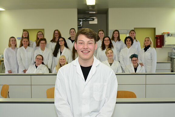 Eine Gruppe junger Menschen in weißen Laborkitteln posiert in einem Unterrichtsraum. Im Vordergrund sitzt ein lächelnder Student im Kittel; dahinter stehen weitere Studierende in Reihen. Die Szene vermittelt Teamarbeit in einem Labor- oder Ausbildungskontext.