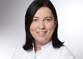 Porträt einer lächelnden Person mit schulterlangem, dunklem Haar im weißen Arztkittel vor neutralem Hintergrund.