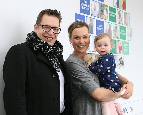Ein Mann und eine Frau lächeln in die Kamera; die Frau hält ein Kleinkind mit Stofftier im Arm. Sie stehen in einem Innenraum, im Hintergrund bunte Poster mit Fotos und Texten.