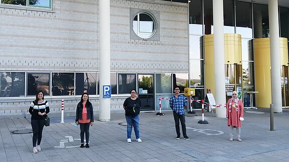 Fünf Menschen stehen mit deutlichem Abstand zueinander draußen vor einem Gebäude in einer Linie. Das Bild zeigt Abstandhalten im öffentlichen Raum.