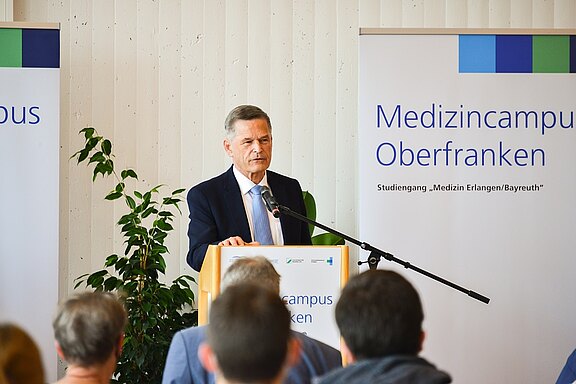 Ein Mann im Anzug spricht an einem Rednerpult mit Mikrofon zu einem kleinen Publikum. Hinter ihm stehen Banner mit der Aufschrift „Medizincampus Oberfranken“ und dem Hinweis auf den Studiengang „Medizin Erlangen/Bayreuth“. Fokus: formelle Ansprache bei einer Hochschulveranstaltung.
