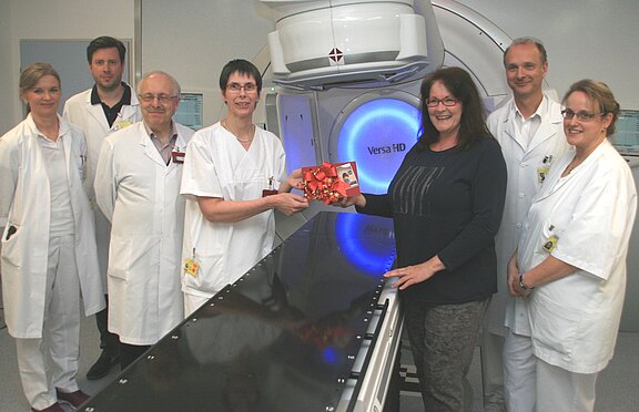 Gruppe von Medizinpersonal in weißen Kitteln steht neben einem großen Strahlentherapiegerät. Eine Mitarbeiterin nimmt von einer Besucherin ein kleines Geschenkpaket entgegen. Alle lächeln; die Szene wirkt wie eine symbolische Übergabe oder Spende im Krankenhaus.