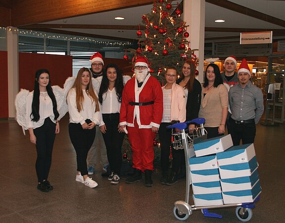 Gruppe von Menschen in Weihnachtskleidung vor einem geschmückten Weihnachtsbaum: ein Weihnachtsmann, zwei Engel und mehrere Personen mit Nikolausmützen. Daneben steht ein Wagen mit gestapelten Kartons.