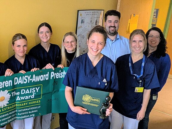 Eine Gruppe von Pflegekräften steht zusammen und hält ein grünes Banner mit der Aufschrift „Wir gratulieren unseren DAISY-Award Preisträgern“. Eine Person zeigt eine Auszeichnungstafel. Feierliche Ehrung für besondere Pflegeleistungen im Krankenhausflur.