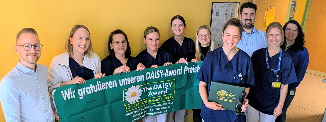 Eine Gruppe von Pflegekräften steht zusammen und hält ein grünes Banner mit der Aufschrift „Wir gratulieren unseren DAISY-Award Preisträgern“. Eine Person zeigt eine Auszeichnungstafel. Feierliche Ehrung für besondere Pflegeleistungen im Krankenhausflur.
