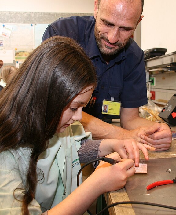 Ein Mädchen lötet konzentriert an einer kleinen Leiterplatte, hält einen Lötkolben. Ein erwachsener Betreuer steht daneben, schaut zu und führt ihre Hand an. Szene in einer Werkstatt oder Unterrichtssituation, die praktische Technikbildung zeigt.