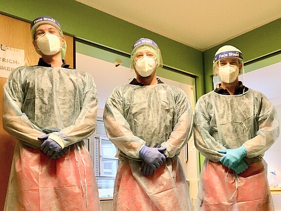 Drei medizinische Fachkräfte stehen nebeneinander in einem Klinikflur. Sie tragen vollständige Schutzkleidung: Gesichtsschilde, Masken, Einwegkittel, Handschuhe und Schutzschürzen. Die Szene vermittelt Infektionsschutz bei der Arbeit.