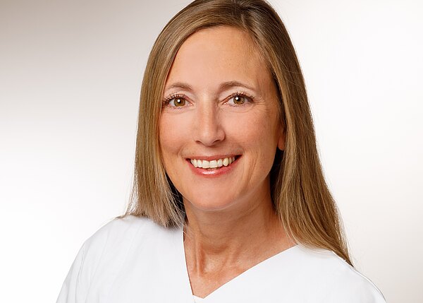 Porträt einer lächelnden Frau in weißer Berufskleidung, vor hellem Hintergrund. Schulterlanges Haar, direkter Blick in die Kamera; wirkt freundlich und professionell.