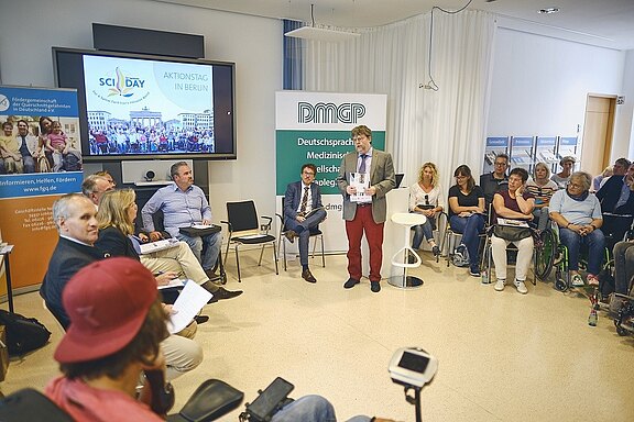 Runder Gesprächskreis bei einer Veranstaltung zum SCI Day in Berlin. Ein Moderator steht mit Unterlagen, umringt von Teilnehmenden, teils im Rollstuhl. Präsentationsbildschirm und Banner der DMGP im Hintergrund. Atmosphäre: Austausch über Querschnittlähmung und Rehabilitation.