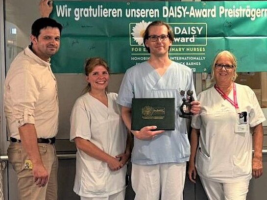 Gruppe von Krankenhausmitarbeitenden posiert in einem Flur unter einem grünen Banner mit der Aufschrift Wir gratulieren unseren DAISY-Award Preisträgern!. In der Mitte hält eine Person in Dienstkleidung eine Trophäe und eine Urkunde. Kolleginnen und Kollegen lächeln daneben, Anerkennung für Pflegeleistungen.