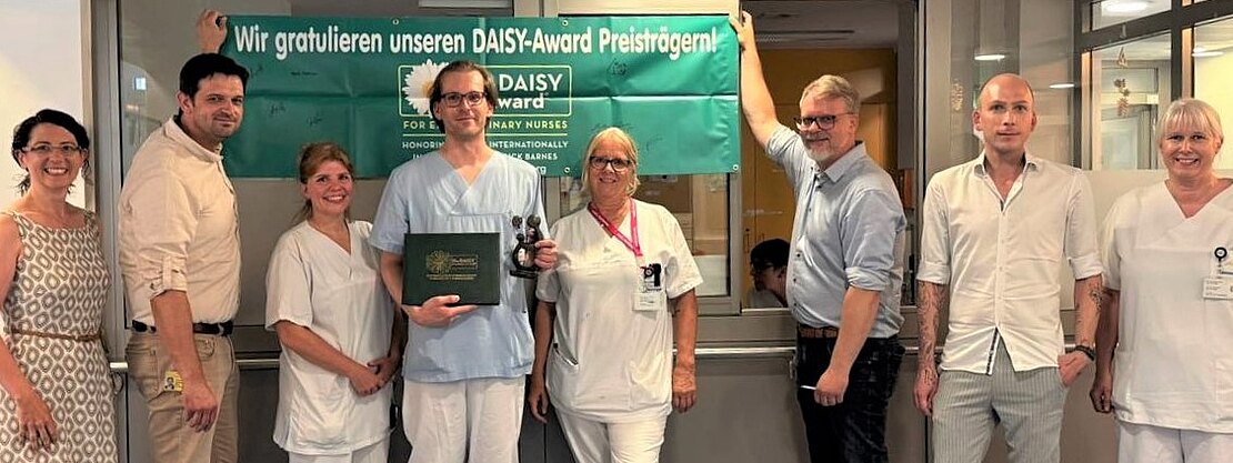 Gruppe von Krankenhausmitarbeitenden posiert in einem Flur unter einem grünen Banner mit der Aufschrift Wir gratulieren unseren DAISY-Award Preisträgern!. In der Mitte hält eine Person in Dienstkleidung eine Trophäe und eine Urkunde. Kolleginnen und Kollegen lächeln daneben, Anerkennung für Pflegeleistungen.