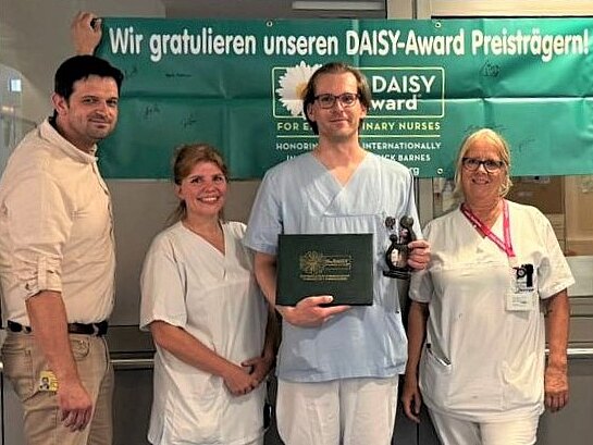 Gruppe von Krankenhausmitarbeitenden posiert in einem Flur unter einem grünen Banner mit der Aufschrift Wir gratulieren unseren DAISY-Award Preisträgern!. In der Mitte hält eine Person in Dienstkleidung eine Trophäe und eine Urkunde. Kolleginnen und Kollegen lächeln daneben, Anerkennung für Pflegeleistungen.