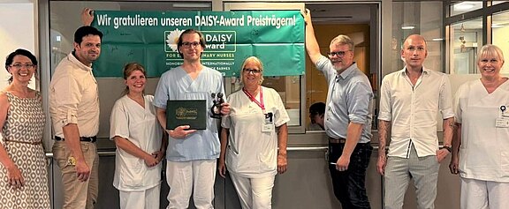 Gruppe von Krankenhausmitarbeitenden posiert in einem Flur unter einem grünen Banner mit der Aufschrift Wir gratulieren unseren DAISY-Award Preisträgern!. In der Mitte hält eine Person in Dienstkleidung eine Trophäe und eine Urkunde. Kolleginnen und Kollegen lächeln daneben, Anerkennung für Pflegeleistungen.