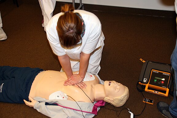 Eine Person führt an einer Reanimationspuppe Herzdruckmassage durch; ein automatisierter externer Defibrillator (AED) ist angeschlossen und liegt daneben.