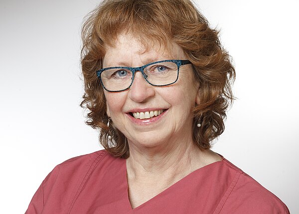 Porträt einer lächelnden Frau mit lockigem rötlichem Haar und blau gerandeter Brille. Sie trägt ein rosa Oberteil vor einem neutralen, hellen Hintergrund.