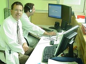 Zwei Ärzte sitzen an einem Schreibtisch in einem Büro. Der Arzt links trägt einen weißen Kittel und lächelt in die Kamera, während der Arzt rechts Kopfhörer trägt und konzentriert auf einen Computerbildschirm schaut. Beide arbeiten am Computer.