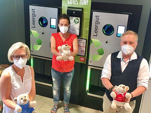 Vor zwei Leergutautomaten in einem Supermarkt stehen drei Erwachsene mit Mund-Nasen-Schutz und halten Teddybären. Auf den Maschinen steht „Leergut“, daneben sind Recycling-Symbole sichtbar.