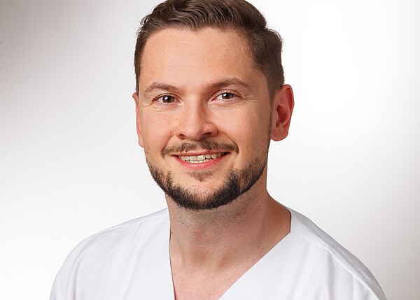 Ein lächelnder Mann mit kurzen, braunen Haaren und einem Bart trägt ein weißes medizinisches Outfit. Er hat einen freundlichen Gesichtsausdruck und steht vor einem hellen, einfarbigen Hintergrund.