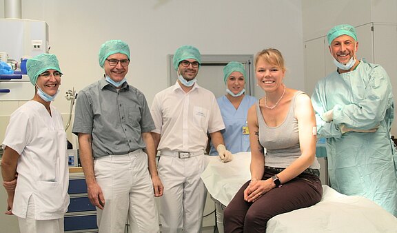 Sechs Personen stehen zusammen in einem medizinischen Raum. Eine Frau sitzt auf einer Liege und lächelt. Alle tragen Schutzkleidung, darunter Hauben und Masken. Die Atmosphäre wirkt freundlich und professionell, was auf eine medizinische Behandlung hindeutet.