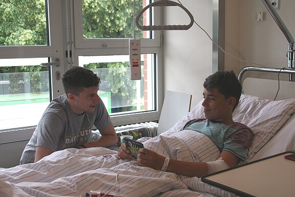 Ein jugendlicher Patient liegt im Krankenhausbett, der Arm bandagiert. Neben ihm sitzt ein Freund, spricht mit ihm und schaut auf ein Heft, das der Patient hält. Die Szene wirkt freundlich und tröstend – ein unterstützender Besuch im Krankenhaus.