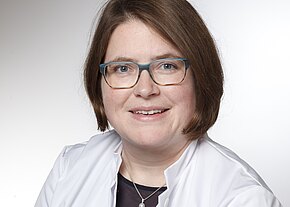 Dr. med. Daniela Senger, MHBA Porträt einer lächelnden Person mit kurzbraunen Haaren und türkisgerandeter Brille, in weißem Kittel vor hellem, neutralem Hintergrund.