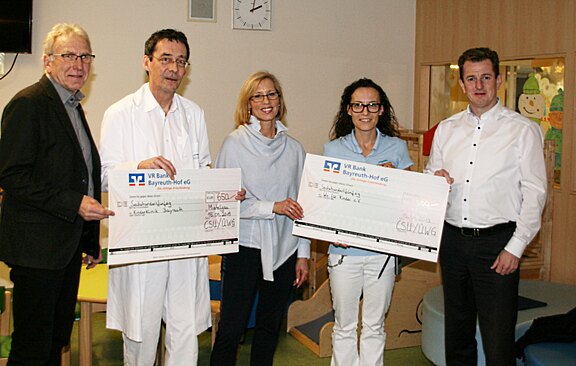 Fünf Erwachsene stehen nebeneinander und präsentieren zwei übergroße Spendenschecks. Ein Mann trägt einen weißen Arztkittel. Die Szene wirkt wie eine Spendenübergabe in einer Kinderklinik.
