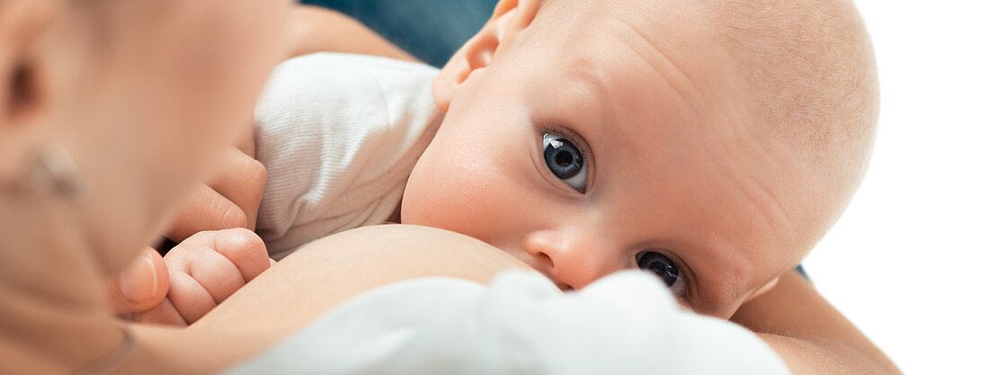 Eine Mutter stillt ihr Baby, das mit großen, neugierigen Augen zur Kamera schaut. Die Intimität des Moments wird durch die enge Umarmung zwischen Mutter und Kind betont. Das Bild vermittelt eine Atmosphäre von Fürsorglichkeit und Verbundenheit.