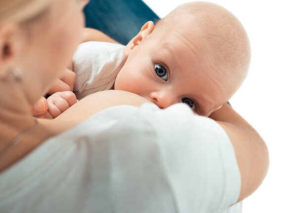 Eine Mutter stillt ihr Baby, das mit großen, neugierigen Augen zur Kamera schaut. Die Intimität des Moments wird durch die enge Umarmung zwischen Mutter und Kind betont. Das Bild vermittelt eine Atmosphäre von Fürsorglichkeit und Verbundenheit.