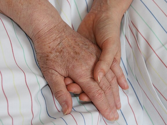 Eine jüngere Hand hält sanft die Hand einer älteren Person auf einem gestreiften Bettlaken. Das Bild vermittelt Trost, Unterstützung und Fürsorge.