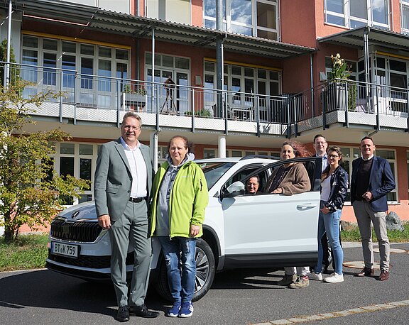 Sieben Erwachsene posieren vor einem mehrstöckigen Gebäude neben einem weißen Auto; eine Person sitzt am Steuer. Gruppenfoto bei Tageslicht.