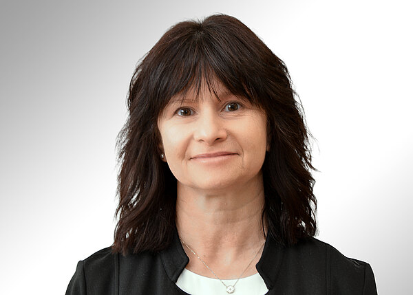 Porträt einer erwachsenen Frau mit mittellangem dunklem Haar, neutralem Gesichtsausdruck und direktem Blick in die Kamera; sie trägt ein dunkles Jackett über einem hellen Oberteil vor hellem Hintergrund.