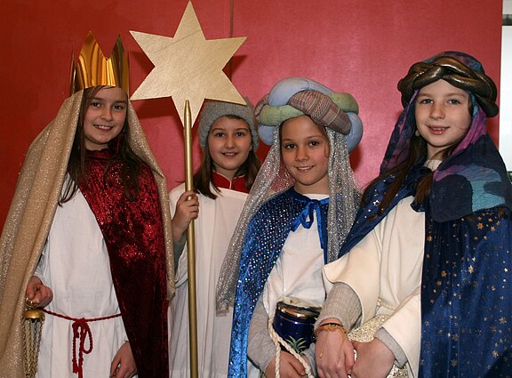 Vier Kinder als Sternsinger/Heilige Drei Könige verkleidet, mit Kronen, Umhängen und Turban; eines hält einen goldenen Stern, ein anderes ein Gefäß. Sie stehen lächelnd nebeneinander vor rotem Hintergrund.