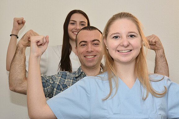 Drei Personen stehen zusammen und zeigen lächelnd ihre starken Arme mit erhobenen Fäusten. Die Person vorne trägt ein hellblaues Oberteil, während die anderen hinten in unterschiedlichen Kleidungen zu sehen sind. Die Gruppe vermittelt ein Gefühl von Stärke und Zusammenhalt.