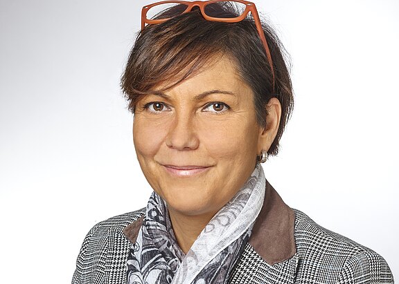 Porträt einer lächelnden Frau vor hellem Hintergrund. Kurzes braunes Haar, orange Brille auf dem Kopf. Sie trägt ein kariertes Sakko mit dunklen Kragenbesätzen und einen hellen gemusterten Schal. Kopf und Schultern im Bild, neutrale, professionelle Darstellung.