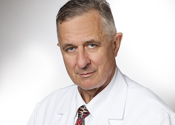 Porträt eines älteren Mannes im weißen Arztkittel und Krawatte vor neutralem Hintergrund, blickt ernst direkt in die Kamera.