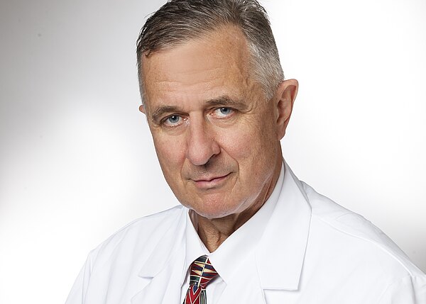 Porträt eines älteren Mannes im weißen Arztkittel und Krawatte vor neutralem Hintergrund, blickt ernst direkt in die Kamera.