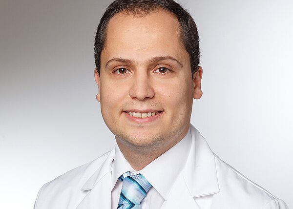 Porträtfoto einer lächelnden Person in weißem Kittel mit blau gestreifter Krawatte vor neutralem Hintergrund.