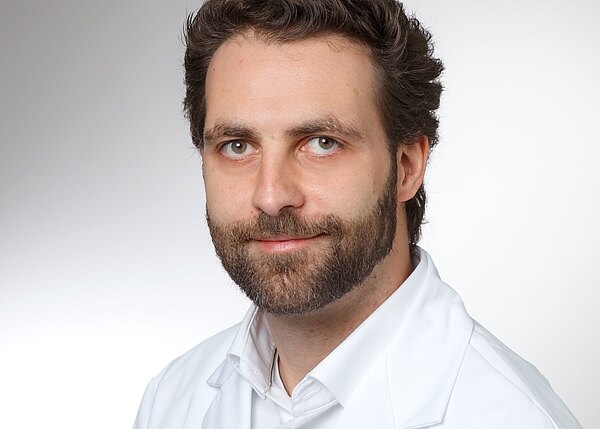 Porträtfoto eines bärtigen Mannes mit dunklem Haar in weißem Kittel, leichtes Lächeln, neutraler Hintergrund, Blick direkt in die Kamera.