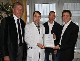 Vier Männer stehen in einem Raum. Einer trägt einen weißen Arztkittel und hält ein Zertifikat in den Händen. Die Männer schauen in die Kamera, einige sind formell gekleidet. Im Hintergrund sind Fenster und eine festliche Dekoration sichtbar.