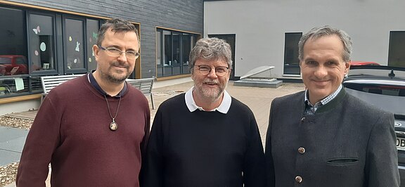 Drei erwachsene Männer stehen nebeneinander und lächeln in die Kamera. Links bordeauxfarbener Pullover mit Kette, in der Mitte schwarzer Pullover über weißem Hemd, rechts graue Jacke mit Stehkragen. Hintergrund: moderner Gebäudeeingang im Freien.