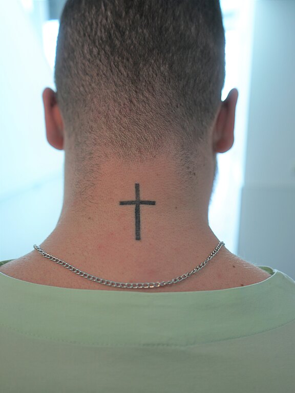 Rückansicht eines kurzhaarigen Kopfes; im Nacken ein schlichtes schwarzes Kreuz-Tattoo. Eine dünne silberne Kette liegt auf den Schultern, Oberteil hellgrün.