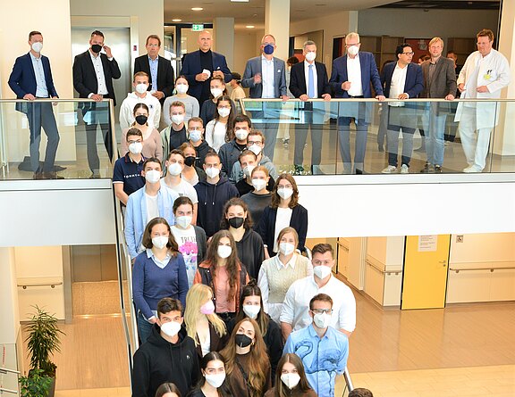 Großes Gruppenfoto in einem hellen Gebäude: Viele Menschen mit Mund-Nasen-Schutz stehen dicht auf einer Treppe, weitere auf der Galerie darüber. Vorne überwiegend Jugendliche, oben Erwachsene in Anzügen und eine Person im weißen Kittel. Pandemie-Situation.